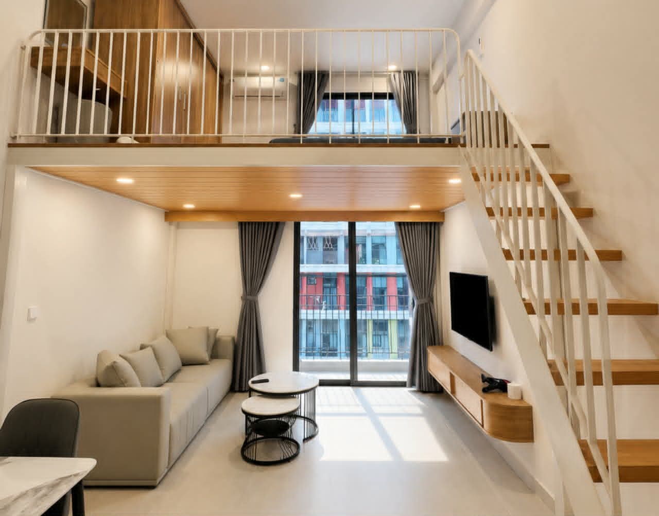 A623 — Toàn cảnh studio gác lửng với cầu thang gỗ, sofa kem và cửa kính ban công